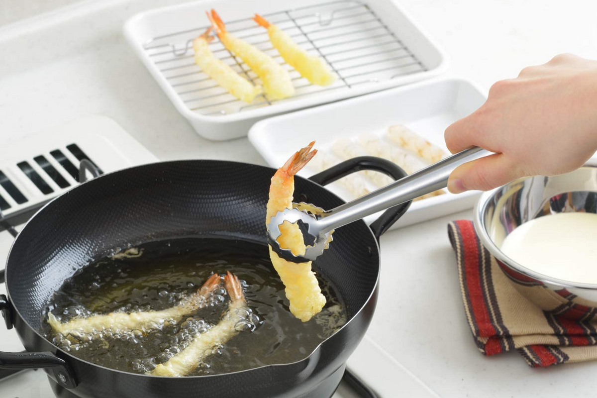 Stainless steel tempura tongs with circular serrated tips gripping a golden fried shrimp tempura over hot oil in black frying pan