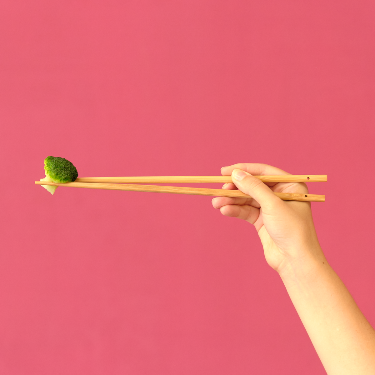 Hand holding Triangular Bamboo Cooking Chopsticks picking up broccoli on pink background
