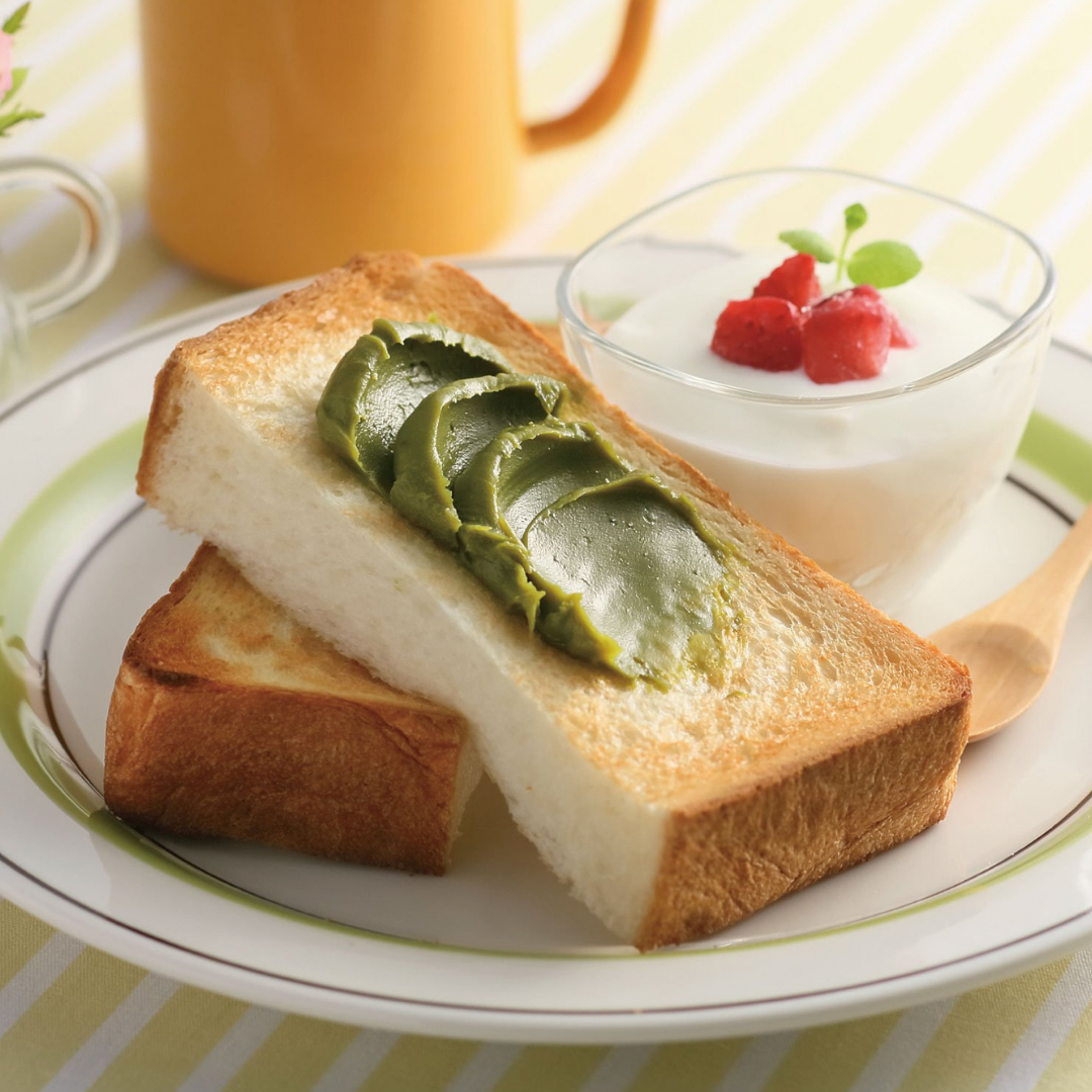 Chikiriya Uji Matcha Milk Jam spread in thick stripes on a slice of white toast on a white plate, served with yogurt topped with strawberries and a yellow mug in the background