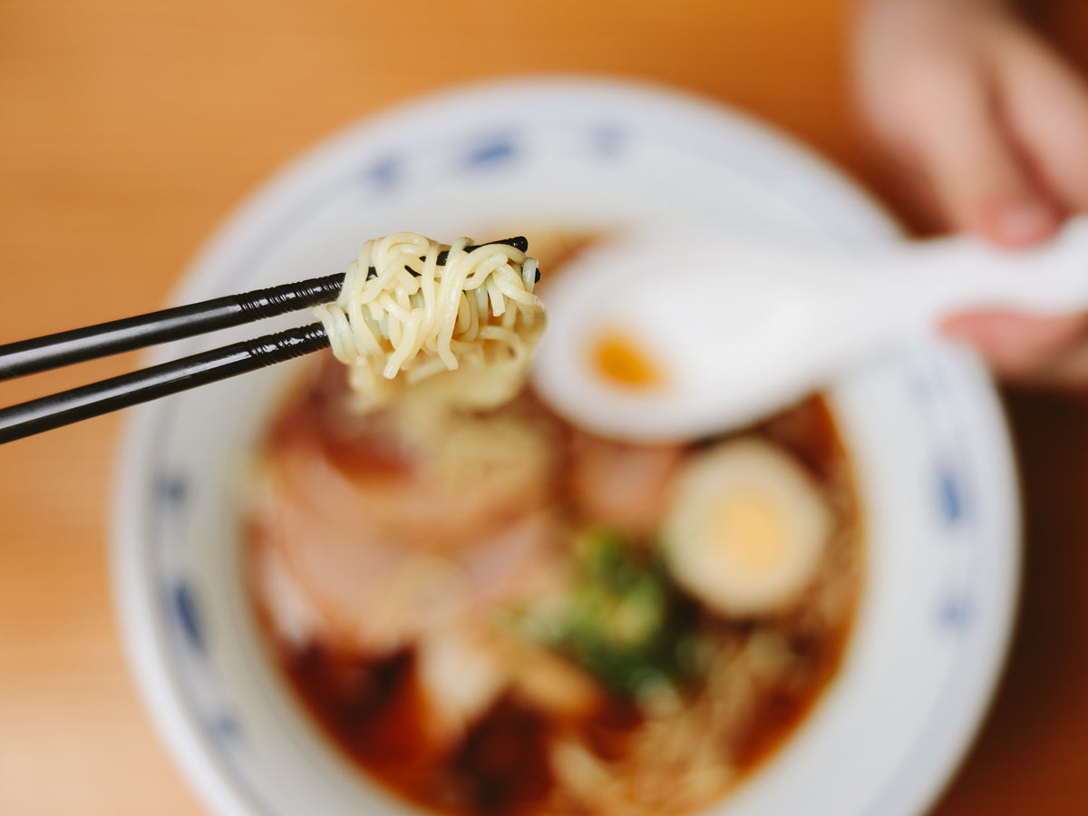 Close-up of Tornado Chopsticks gripping ramen noodles above a bowl with soft-boiled egg and chashu pork