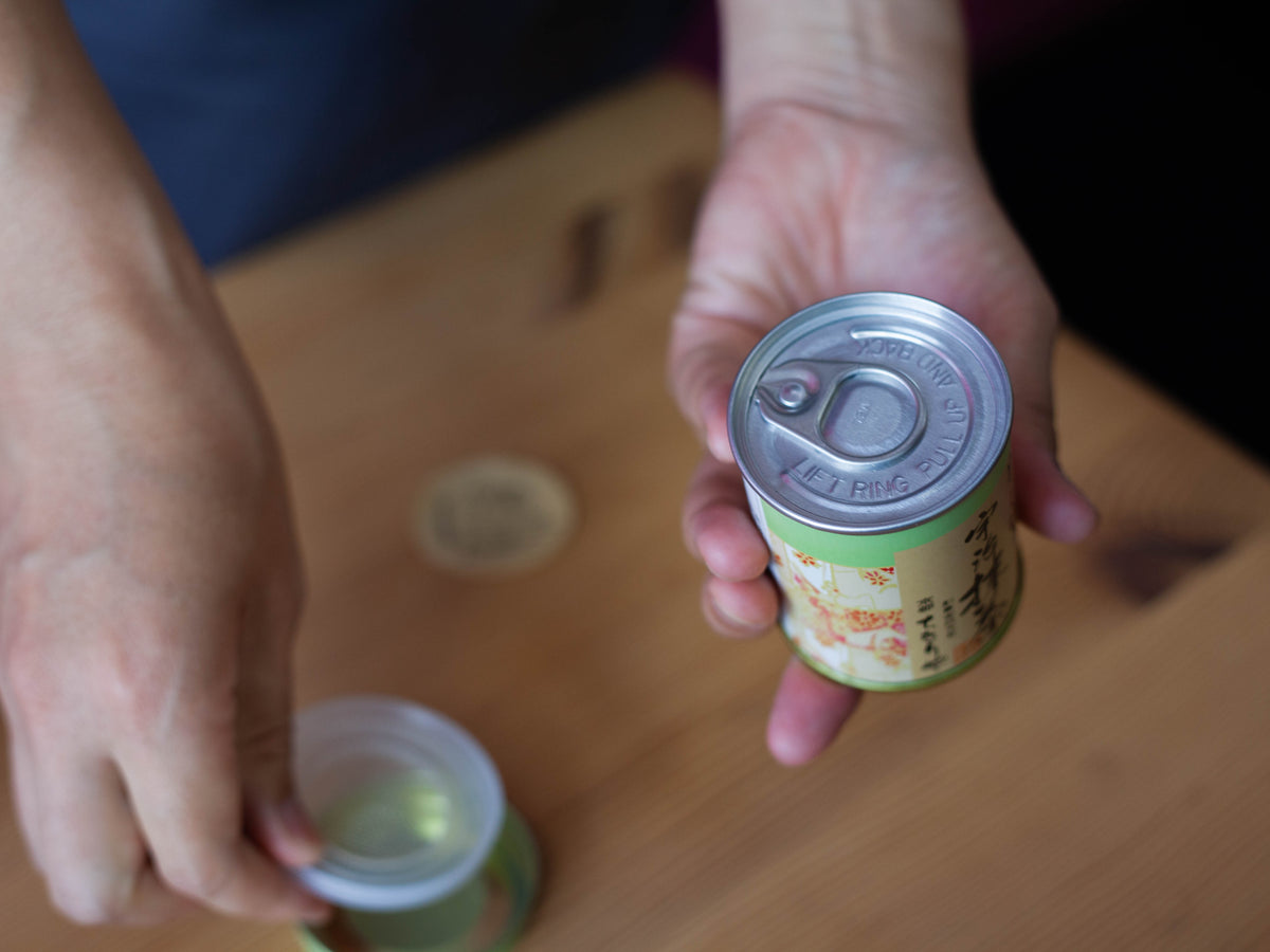 Hands holding the Chikiriya Uji Matcha Green Tea Powder tin can upside down, pulling the ring tab to open the sealed bottom, on a wooden table