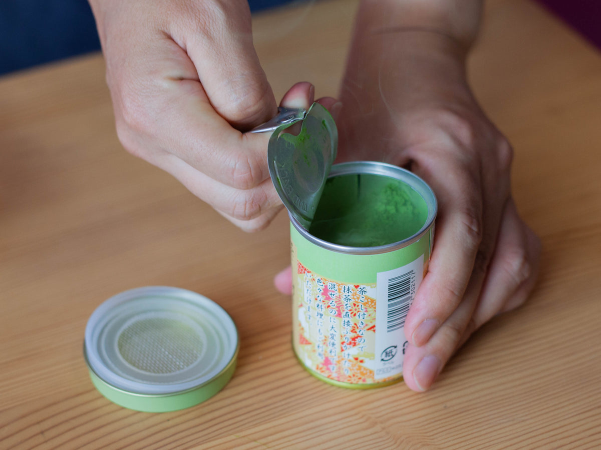 Hands peeling open the pull-tab lid of the Chikiriya Uji Matcha Green Tea Powder tin can, revealing the vivid green matcha powder inside, on a wooden table