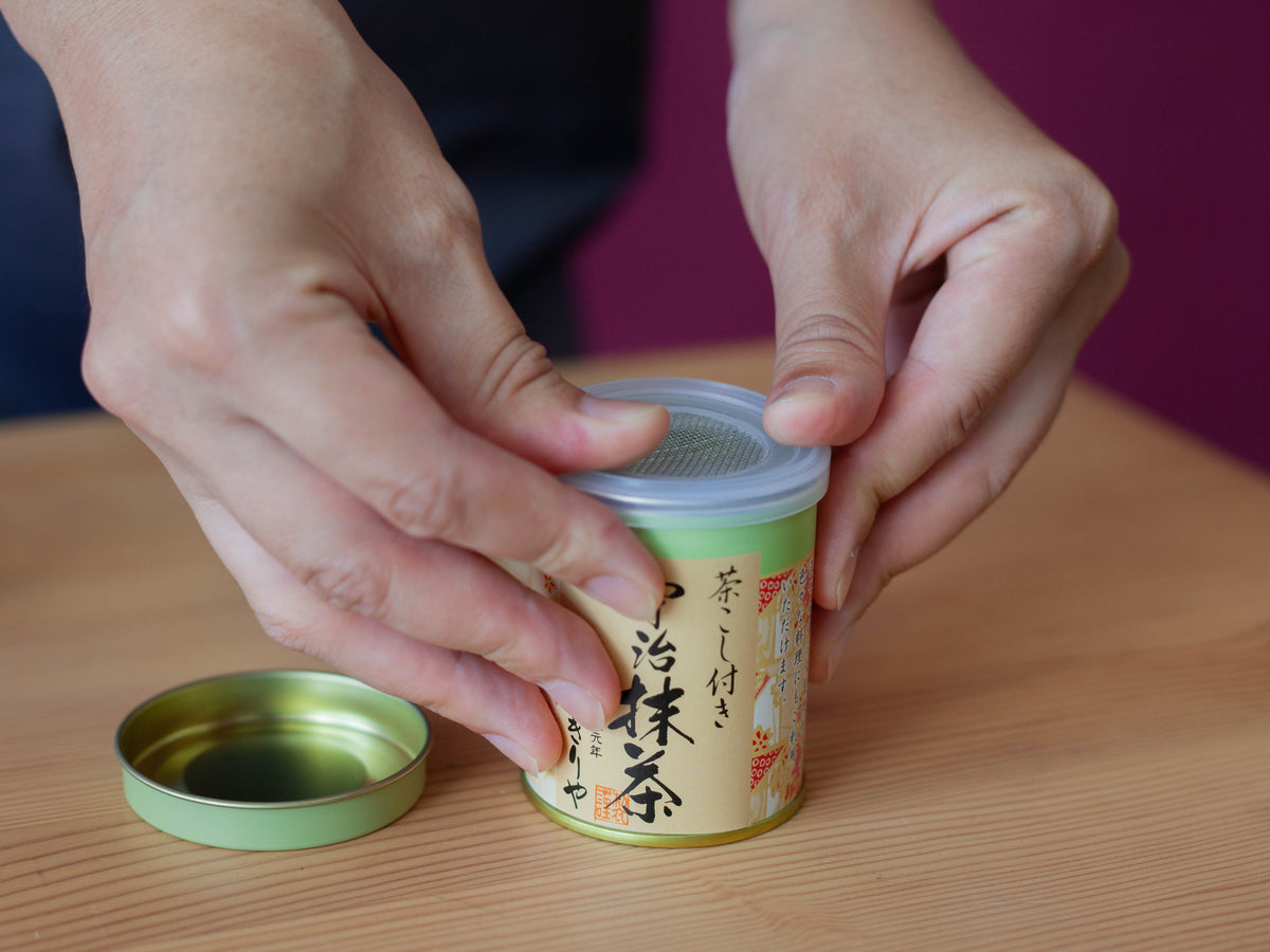Hands peeling open the pull-tab lid of the Chikiriya Uji Matcha Green Tea Powder tin can, revealing the vivid green matcha powder inside, on a wooden table
