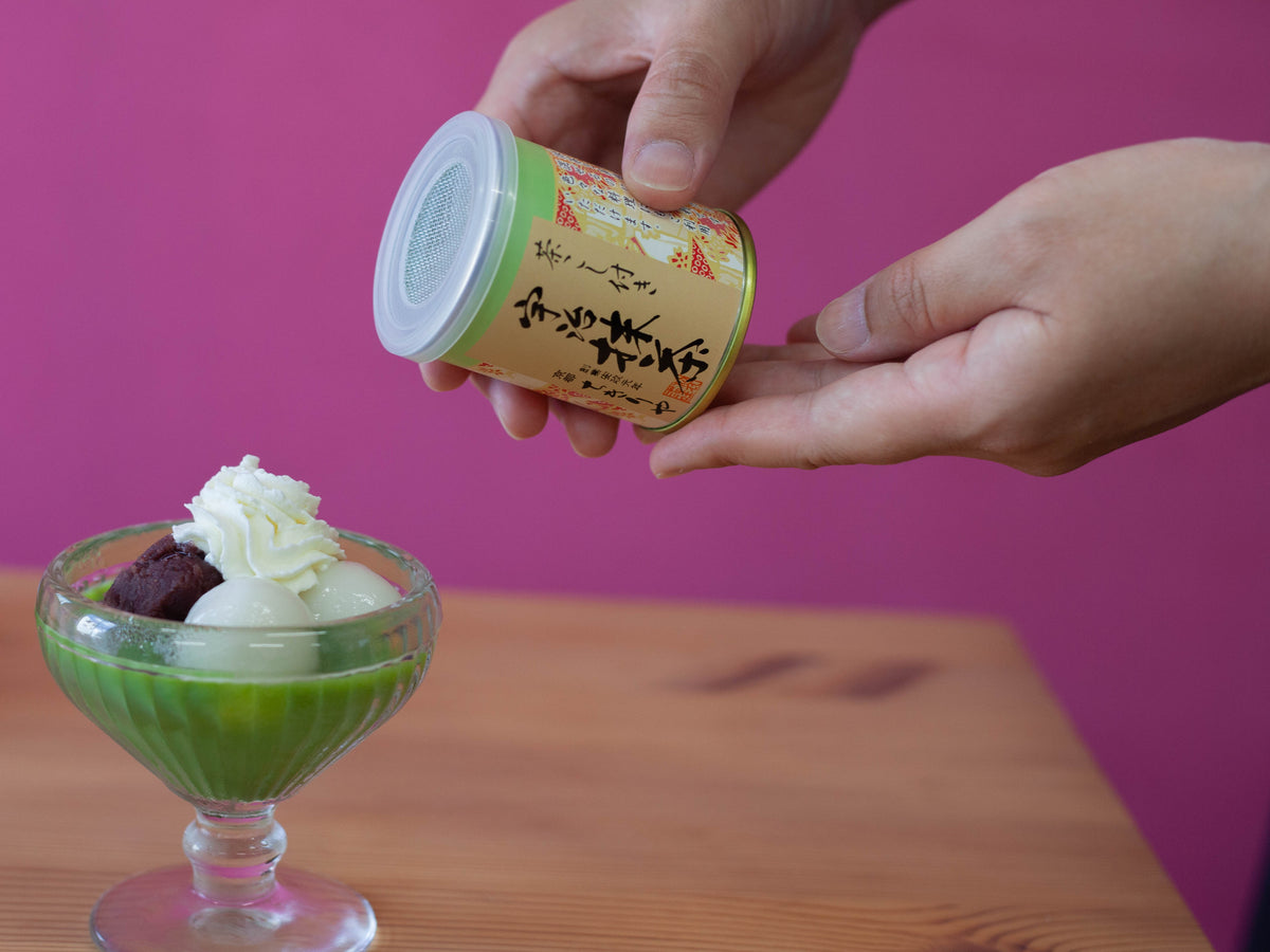 Sifting Chikiriya Uji Matcha Green Tea Powder over a Matcha parfait in a glass leaf-shaped bowl on a ceramic plate, using the built-in sifter lid
